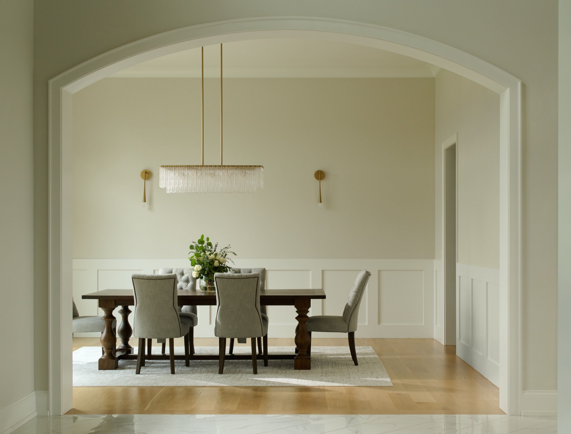 The formal dining room framed by a graceful arched opening, with a linear glass chandelier and upholstered seating.