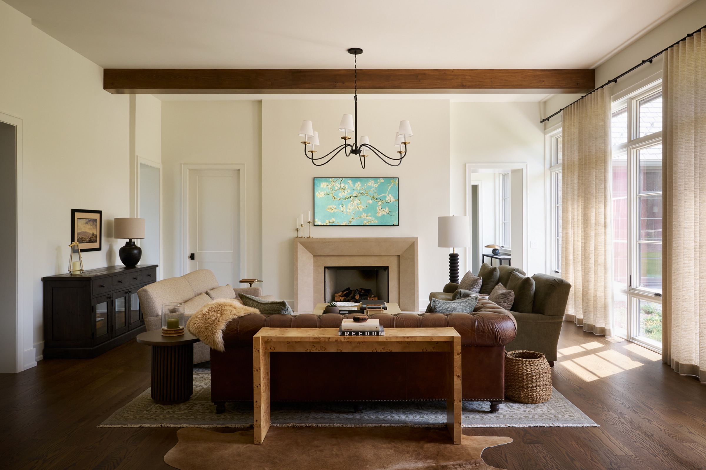 Spacious and sun-filled, this open-concept living room in the main body of the home features a marble fireplace centered in the room, an exposed wooden beam ceiling, floor-to-ceiling windows, and French doors that reveal the beautifully landscaped backyard.