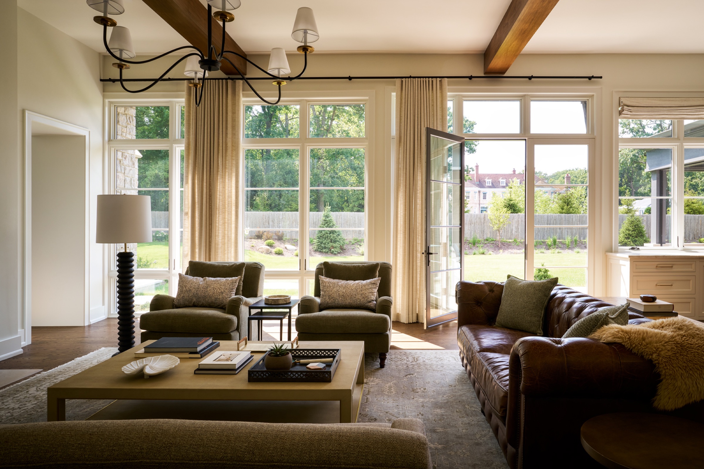 A sun-filled, open-concept living room and kitchen area in the main body of the home, featuring expansive transom and tall casement windows, exposed wooden beam ceilings, and wide-plank hardwood flooring.