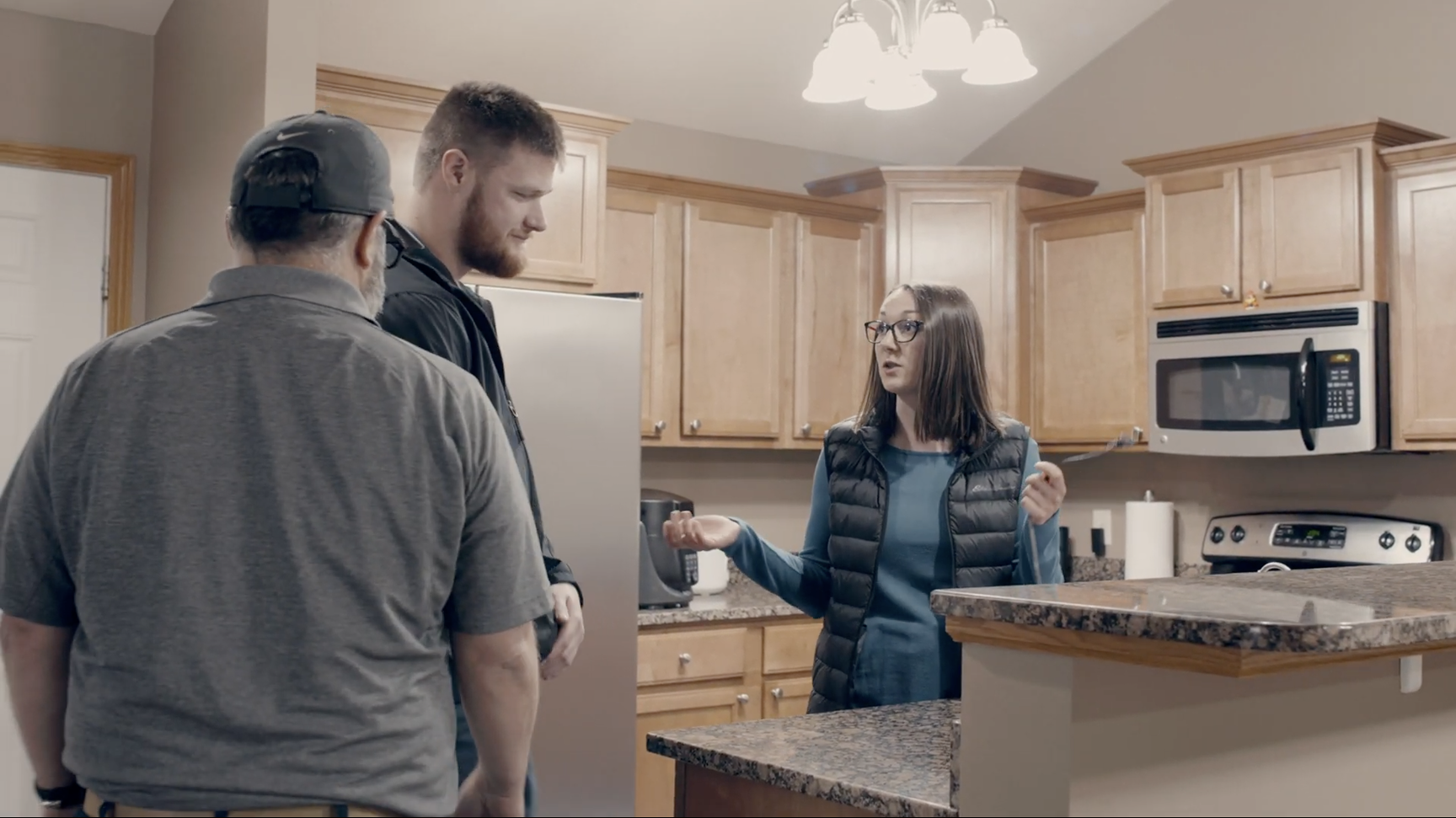 Three people having a conversation in a kitchen with wooden cabinets and granite countertops.