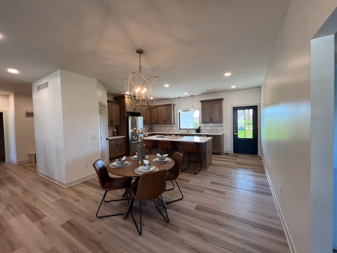 Modern kitchen with wooden cabinets, a center island with brown bar stools, round dining table set for four with brown chairs, and a black door leading outside.