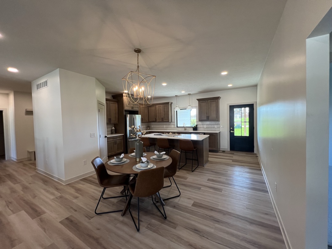 Modern kitchen with wooden cabinets, a center island with brown bar stools, round dining table set for four with brown chairs, and a black door leading outside.