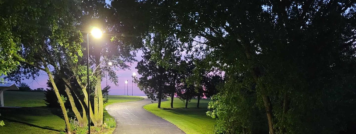 Curved paved path illuminated by streetlights at dusk with trees and grassy areas on both sides.