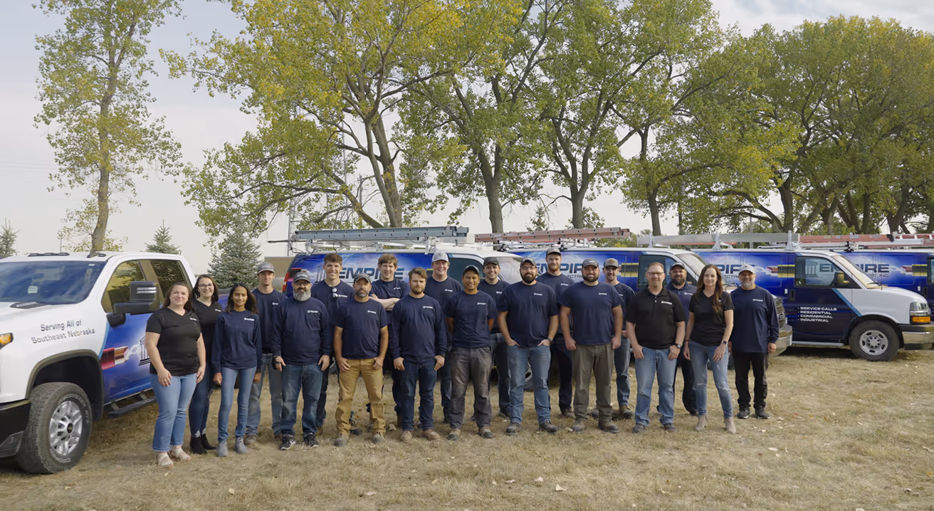 Empire Electric team standing in a line outdoors on grass in front of work vans with company logos and ladders on top, with trees in the background.