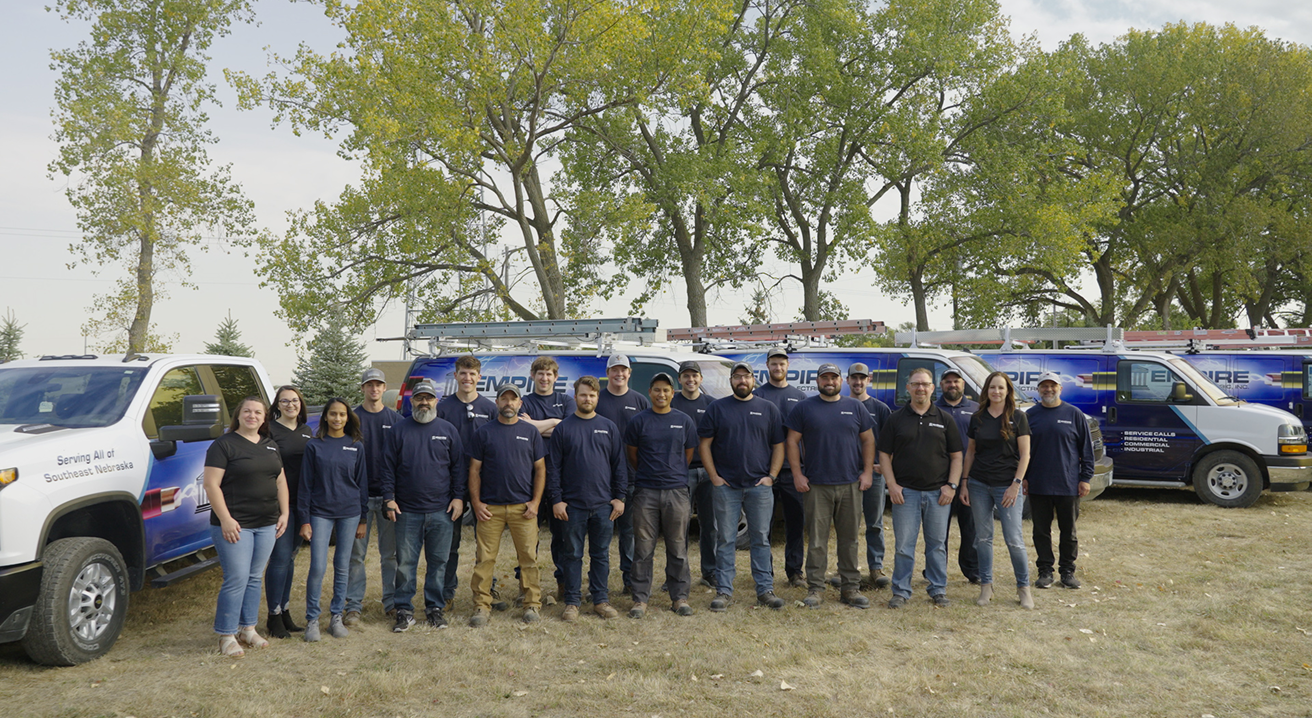 Empire Electric team standing in a line outdoors on grass in front of work vans with company logos and ladders on top, with trees in the background.