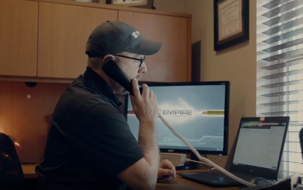 Man wearing a cap and glasses talking on a corded phone while sitting at a desk with a laptop and a monitor displaying 'EMPIRE'.
