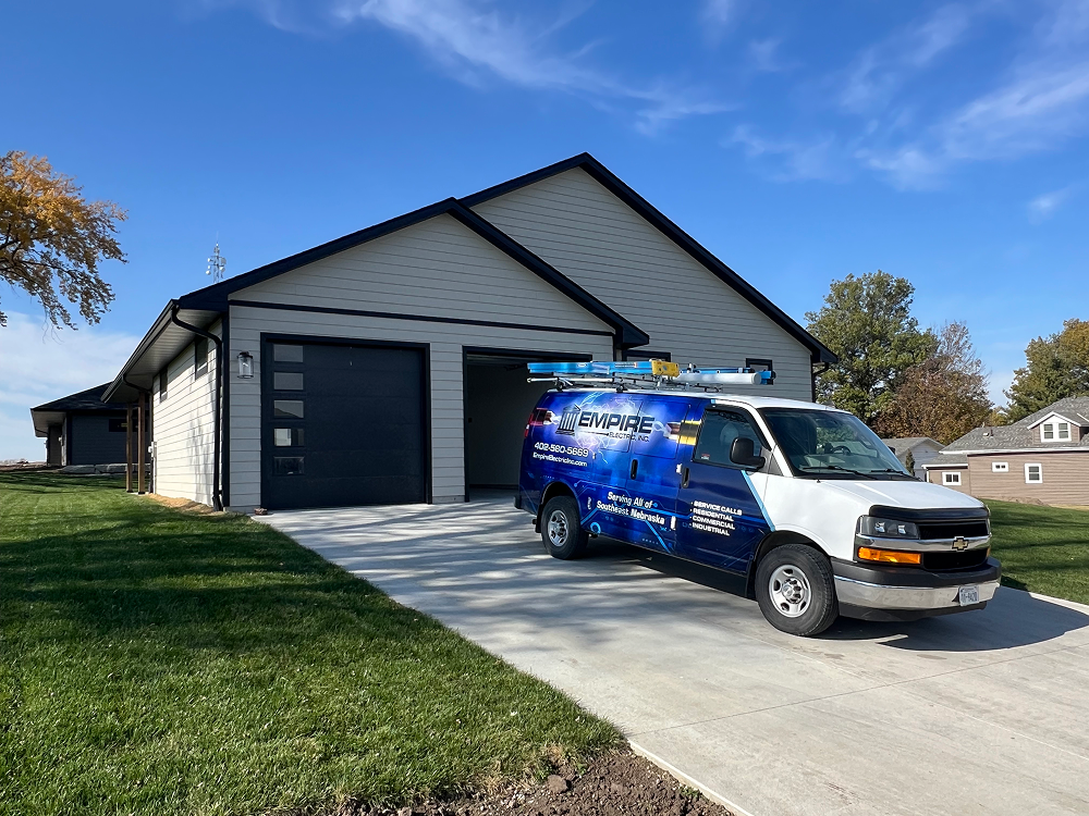 Blue and white Empire Electric work van parked on the driveway of a modern beige house with black trim and garage doors.