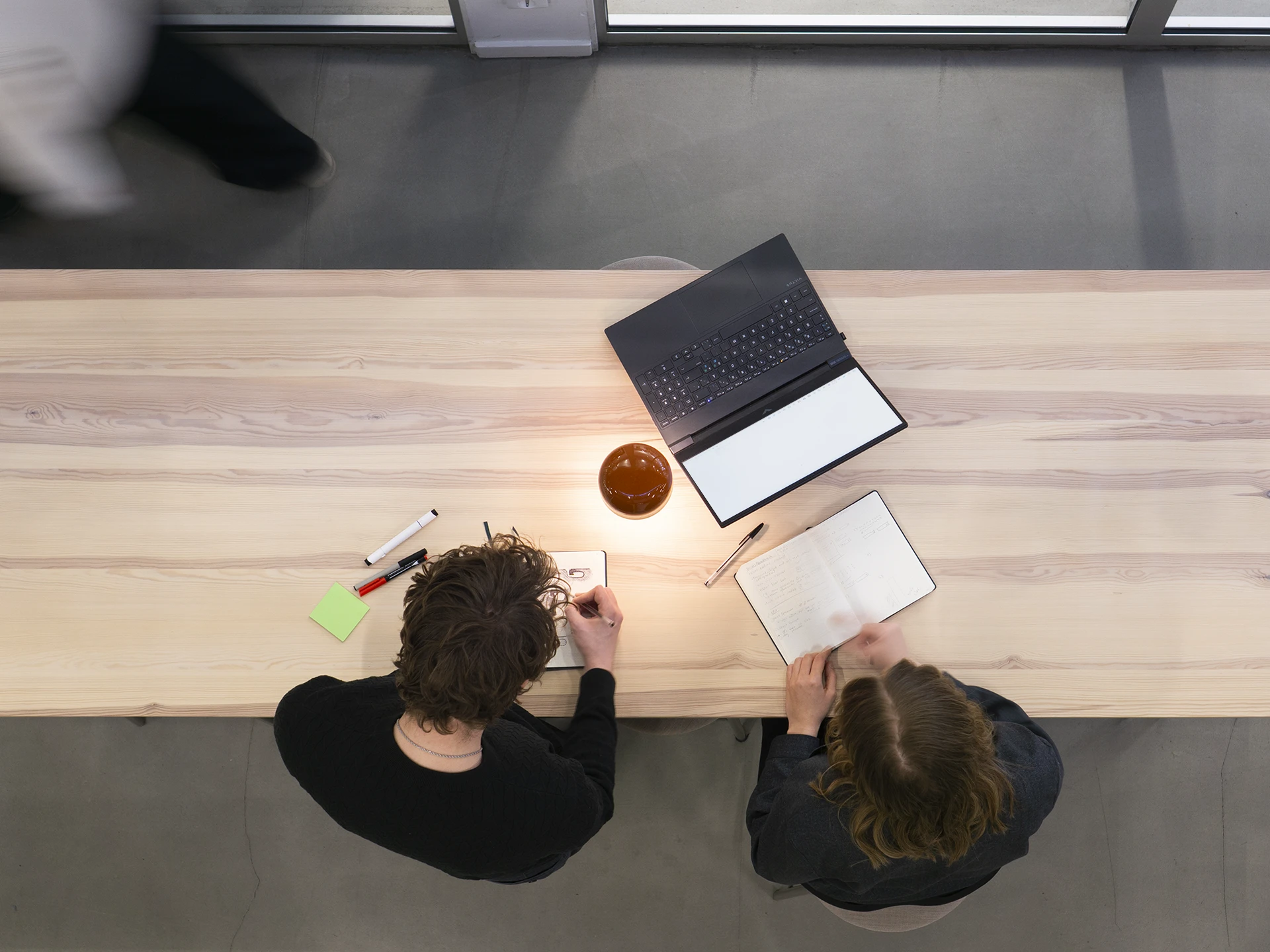 Overhead view of colleagues collaborating at a long table with a blurred figure walking in the background.