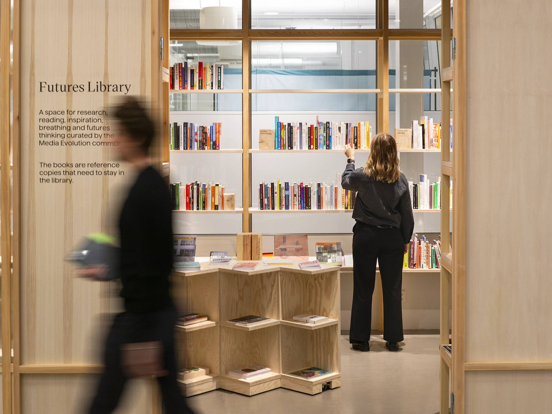 Designer reaching for a book in the studio library while a colleague walks past blurred in the foreground.