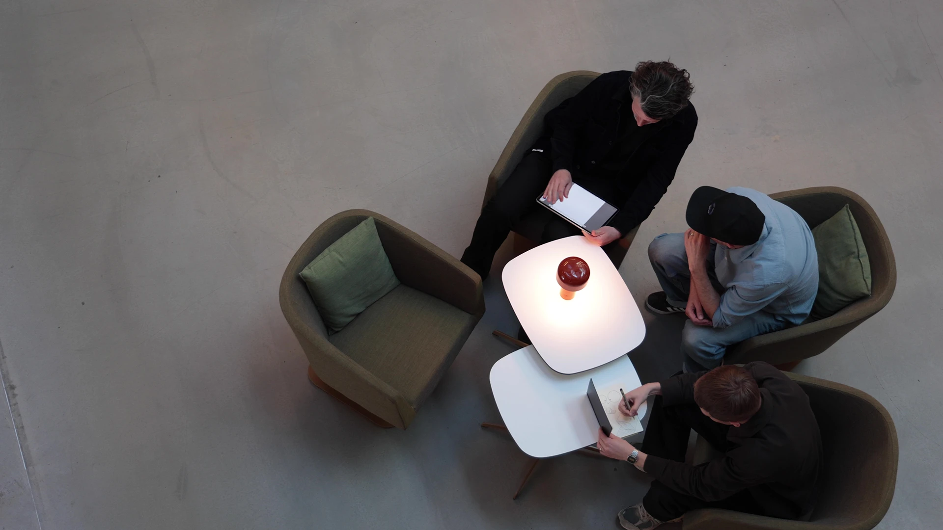 Three people sitting on armchairs around two small tables, one person writing on a notepad, another holding a tablet.