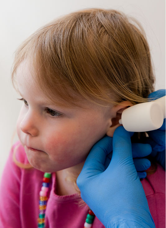 Child in pink shirt gets ear checked with white device by person wearing blue gloves.