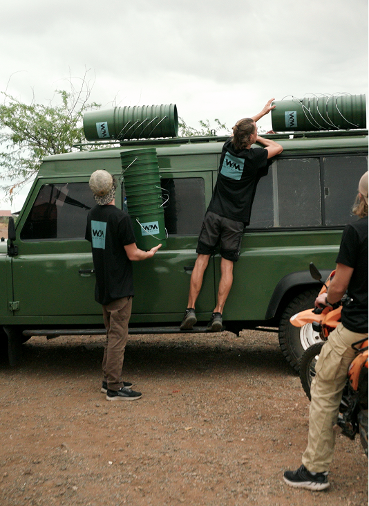 Three people load green buckets onto the roof of a green off-road vehicle in a dirt area.