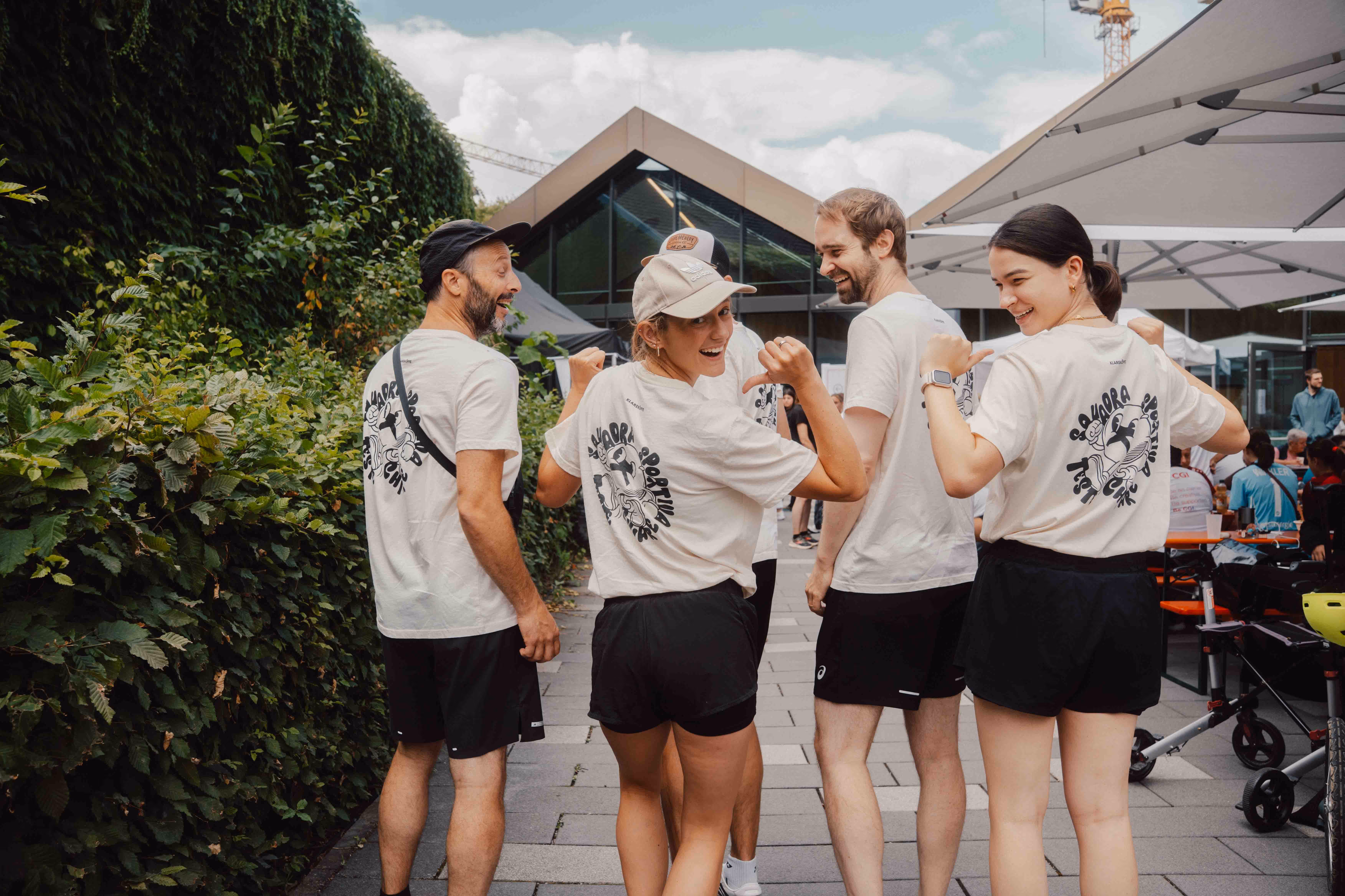 Vier lächelnde Personen mit weißen T-Shirts und schwarzen Shorts zeigen die Rückseite ihrer Shirts draußen – ready für den Spendenlauf.