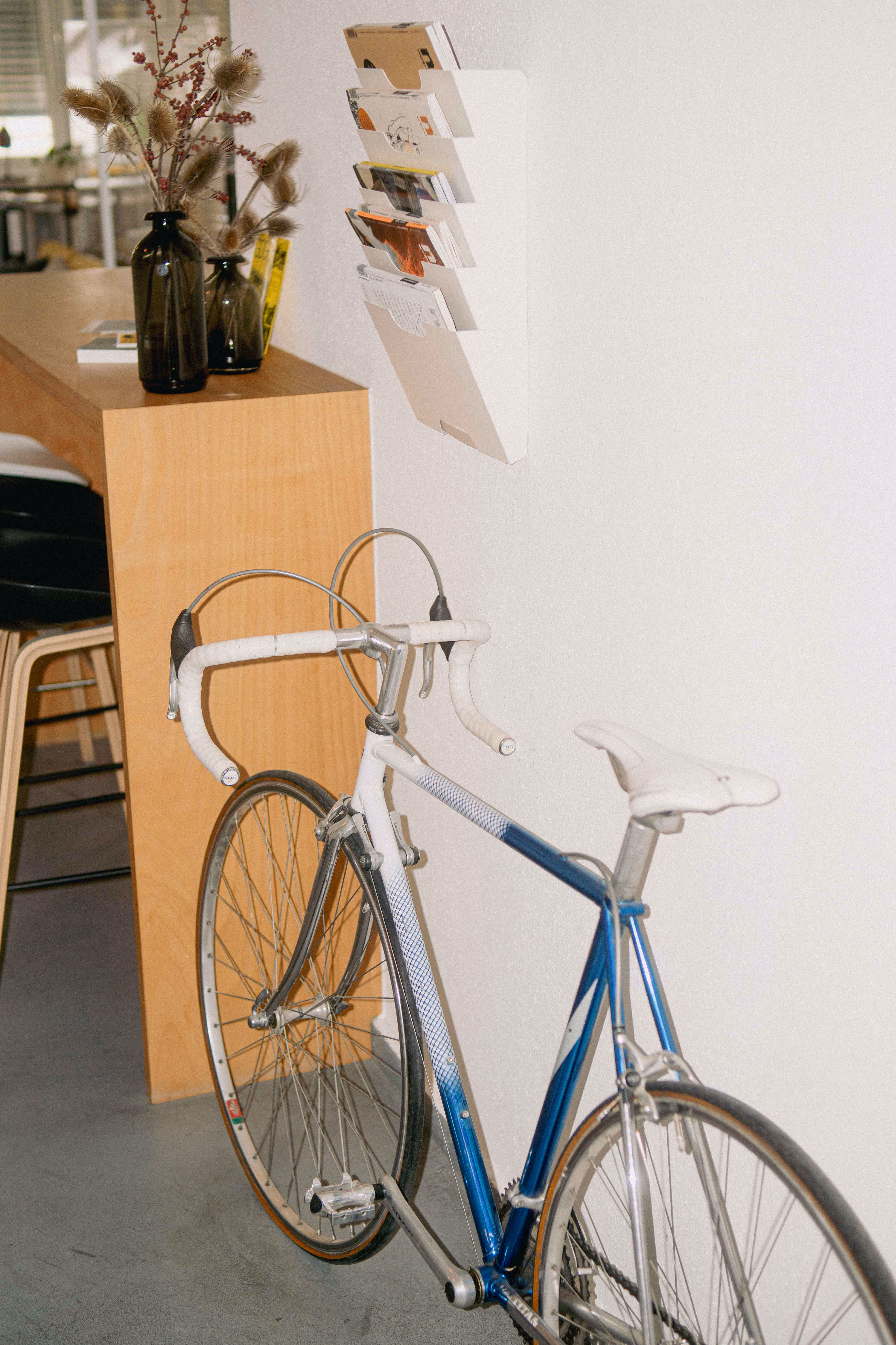 Blau-weißes Rennrad lehnt an weißer Wand neben Holztheke mit zwei Vasen und Wandzeitschriftenhalter im Office von KLAREKÖPFE.