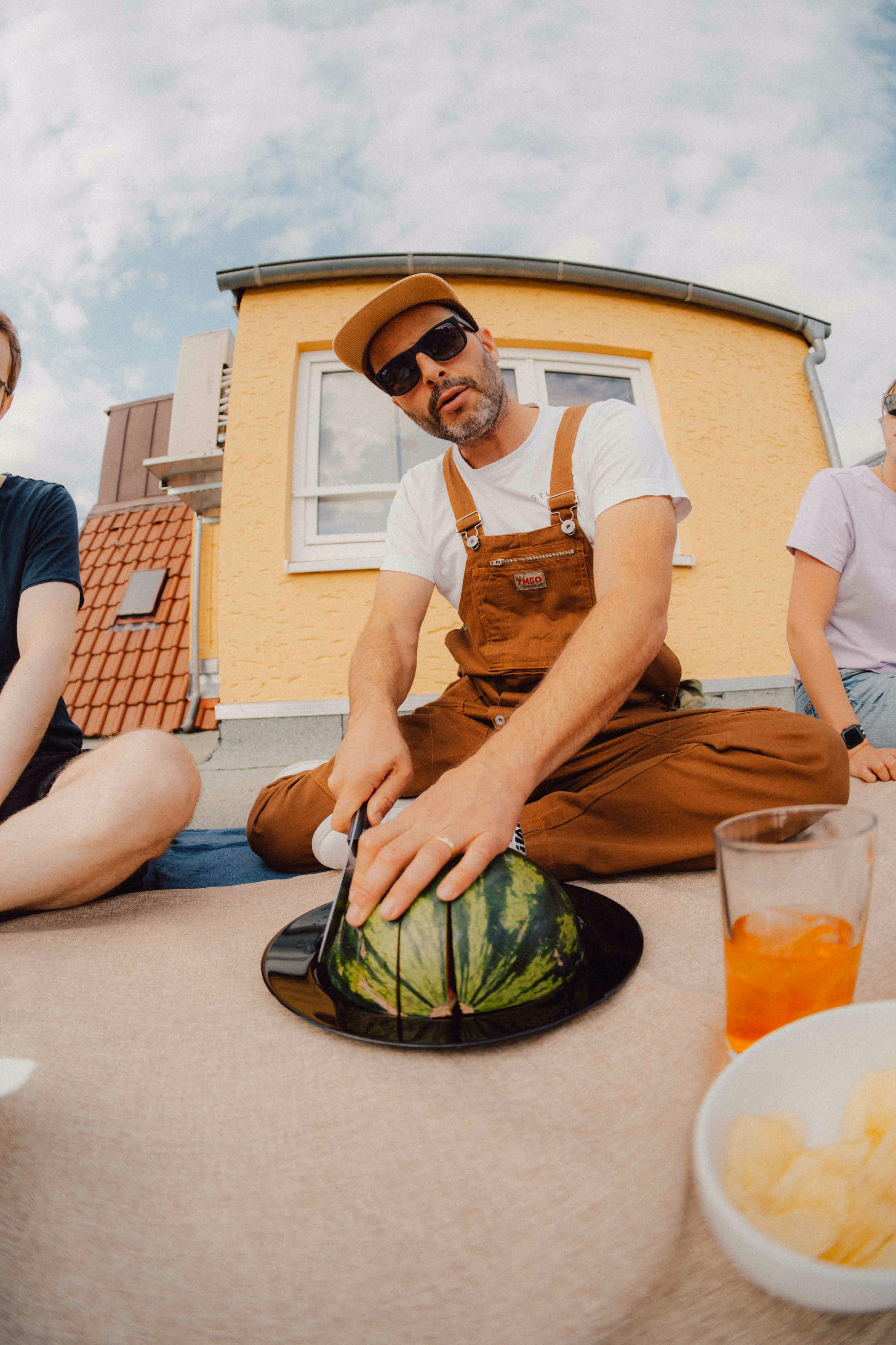 Mann in brauner Latzhose, mit Sonnenbrille und Cap schneidet eine Wassermelone auf einem Dach, umgeben von Freunden.