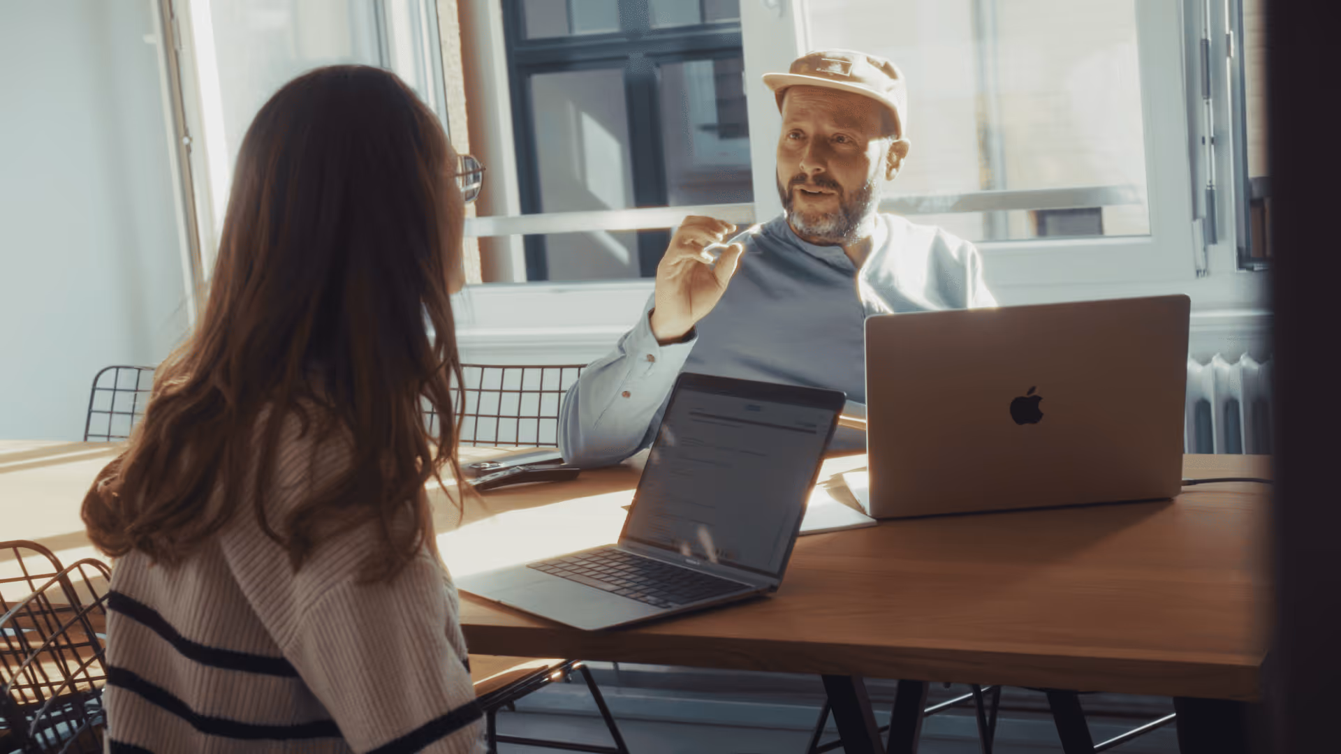 Mann mit Kappe erklärt Frau am Tisch mit geöffneten Laptops vor Fenster im Büro.