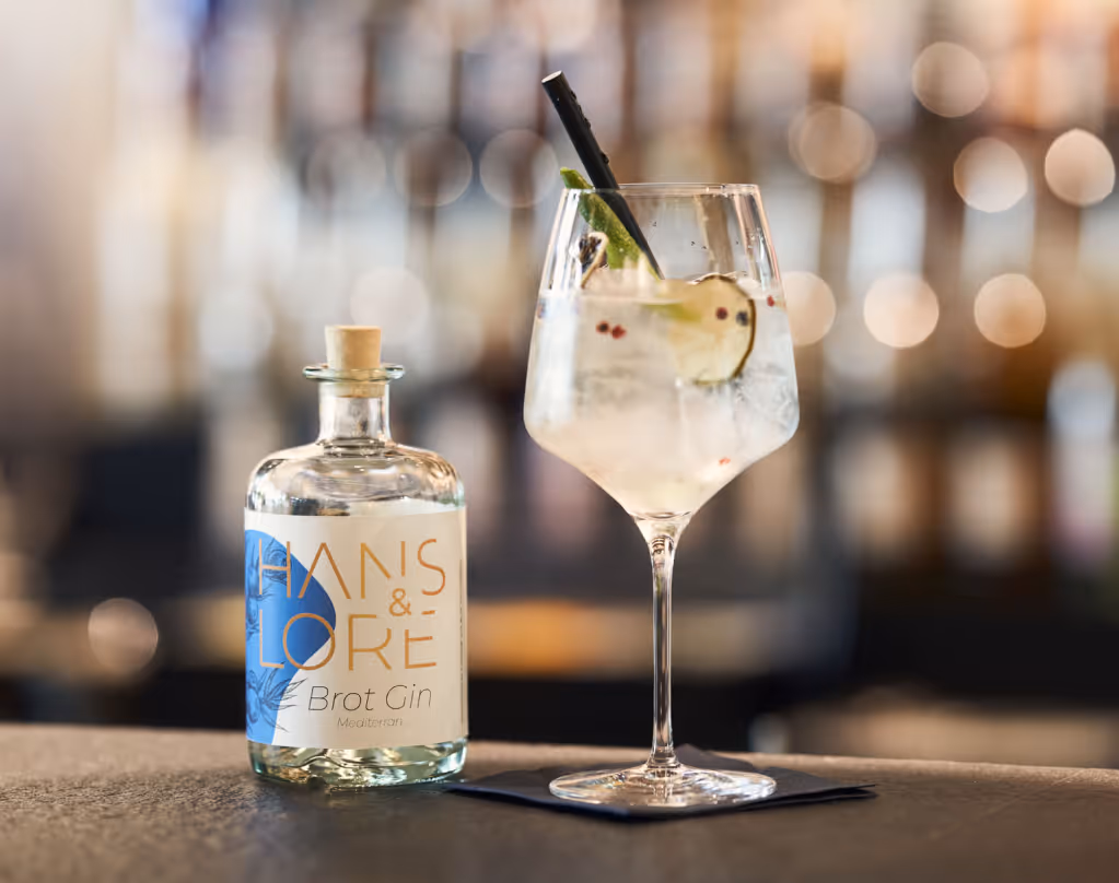 Flasche der Spirituose von "Hans & Lore" neben einem mit Gurke und Eiswürfeln gefüllten Gin-Glas mit Strohhalm.