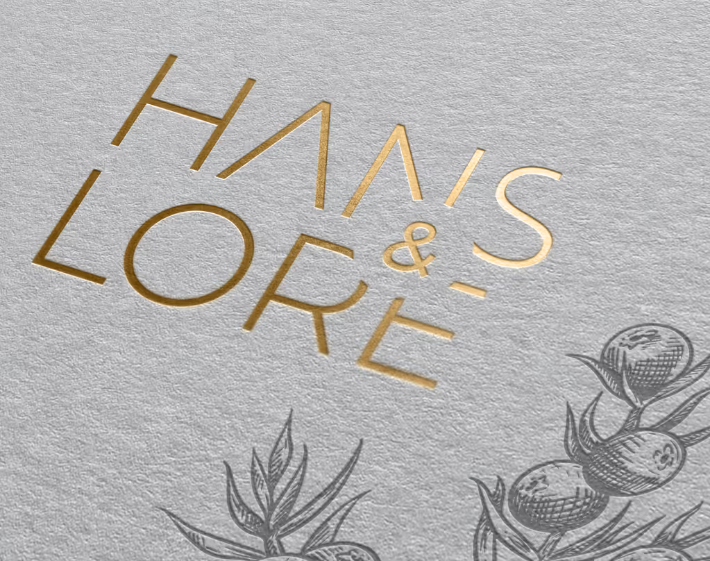 Goldgeprägter Schriftzug 'HANS & LORE' auf grauem Naturpapier mit Zeichnungen von Wacholderbeeren unten rechts.