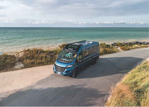 Blue camper van parked on a coastal road near the ocean with cloudy sky.