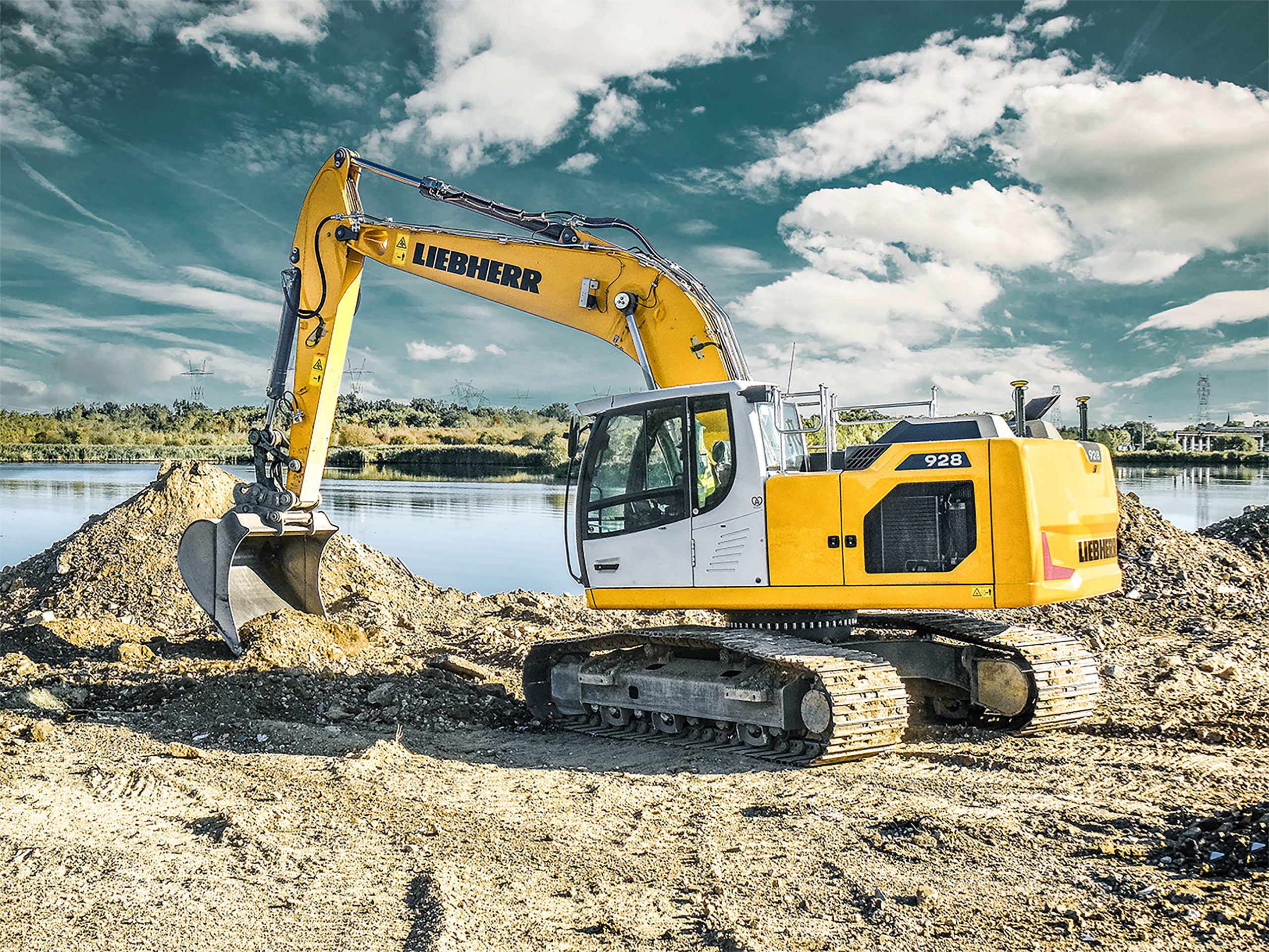 Yellow Liebherr 928 excavator digging soil near a water body under a partly cloudy sky.