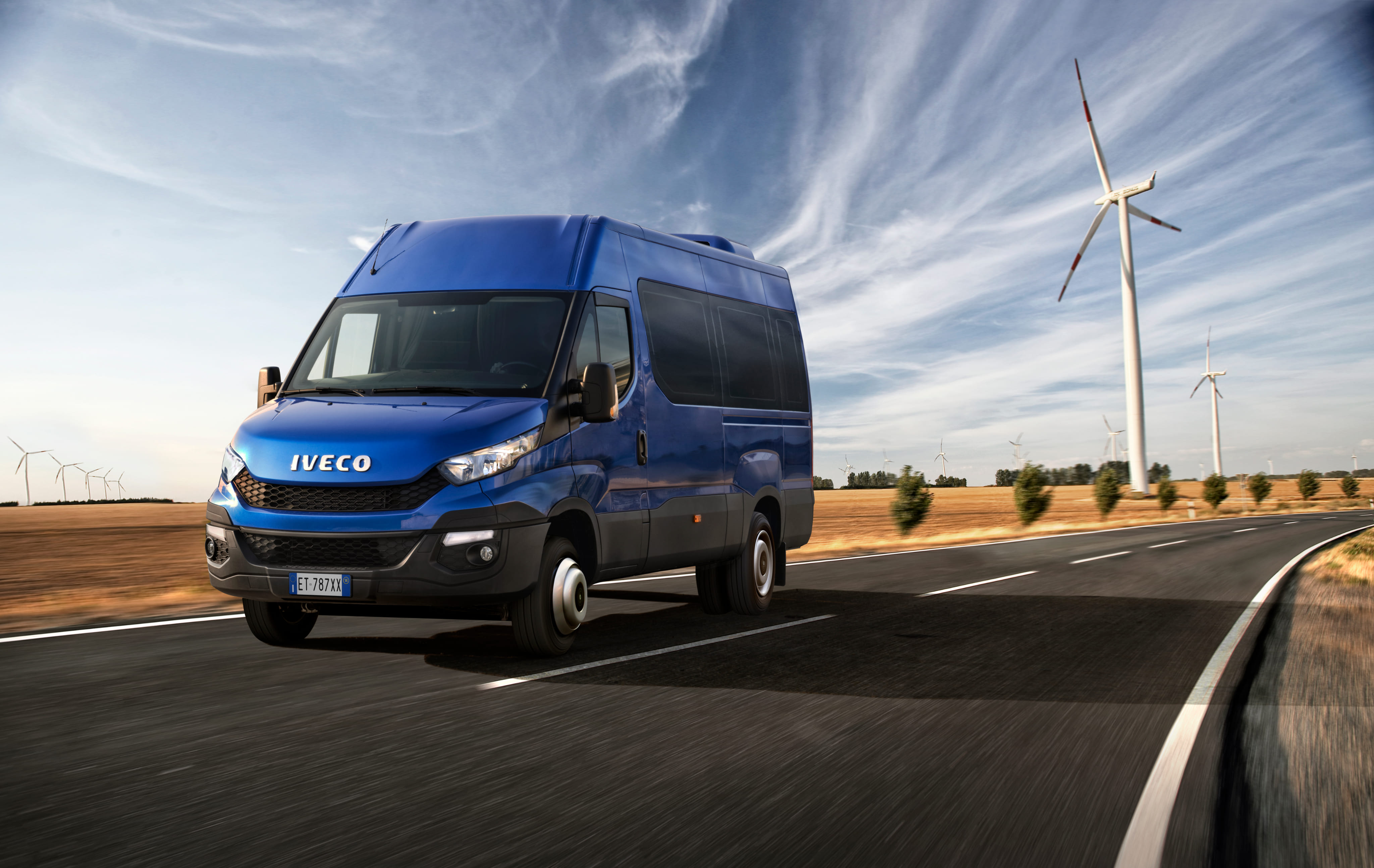 Blue Iveco van driving on curved road with wind turbines and dry fields under a partly cloudy sky.