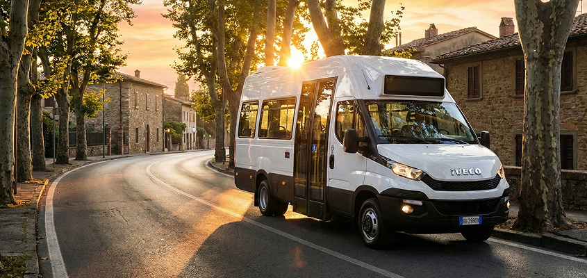 White Iveco van parked on a sunlit curved road with stone houses and tall trees at sunset.