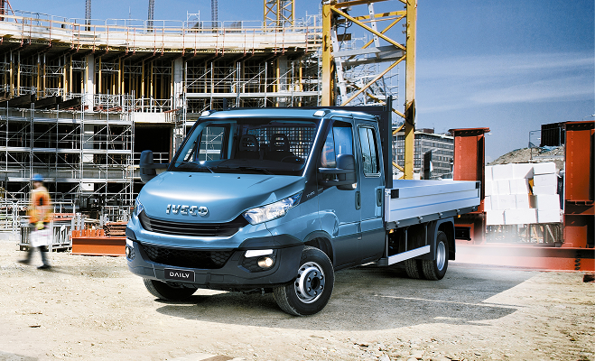 Blue Iveco Daily crew cab flatbed truck parked at a construction site with scaffolding in background.