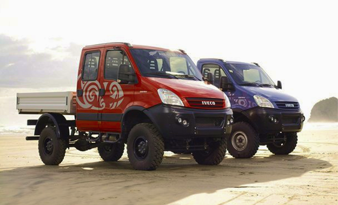 Two Iveco off-road trucks, one red with white design, one blue, parked side by side on a beach.