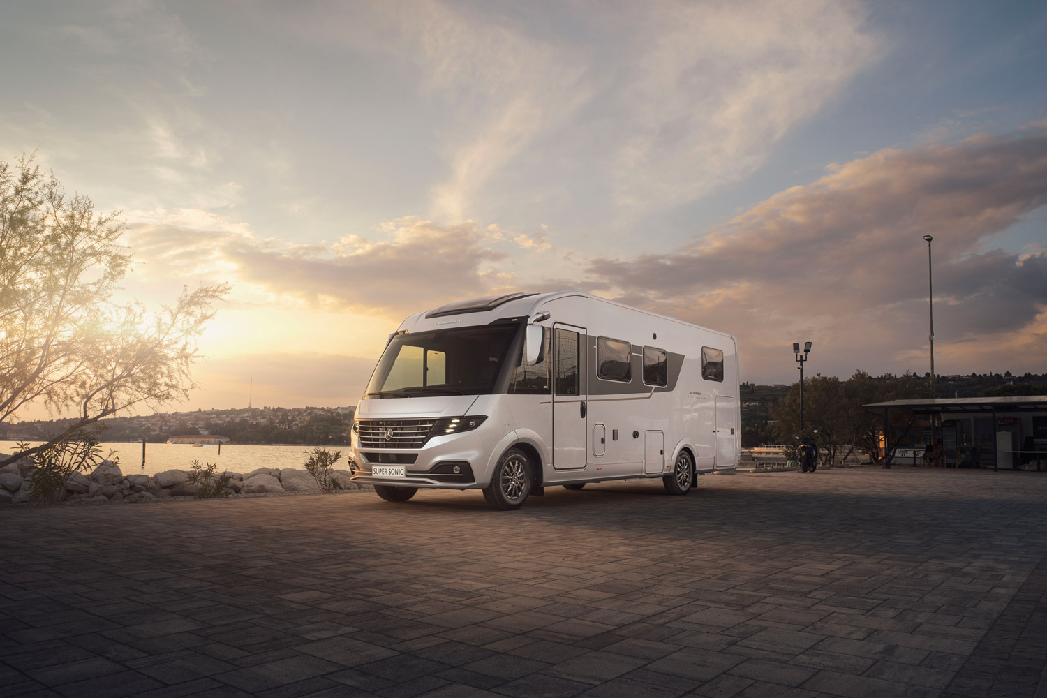 White luxury camper van parked on a paved area near water at sunset with a partly cloudy sky.