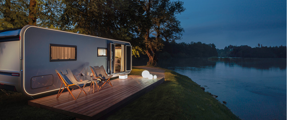 Modern camper trailer with deck, chairs, and glowing lights beside calm lake at dusk.