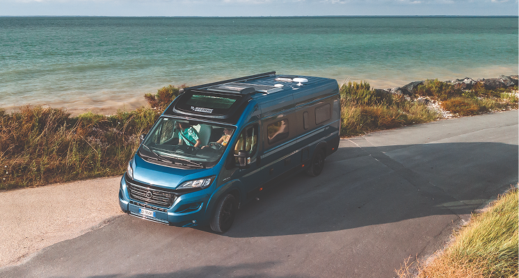 Blue camper van parked on coastal road beside sea and greenery under a partly cloudy sky.