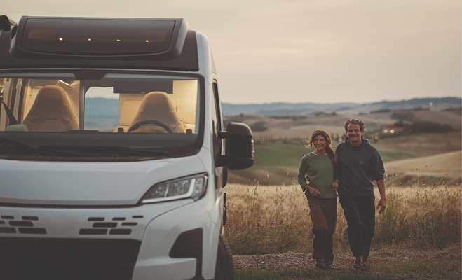 Couple walking arm in arm near a white campervan in a grassy countryside at sunset.