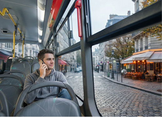 Young man in a gray hoodie sitting alone on a bus, talking on phone, city street visible outside.