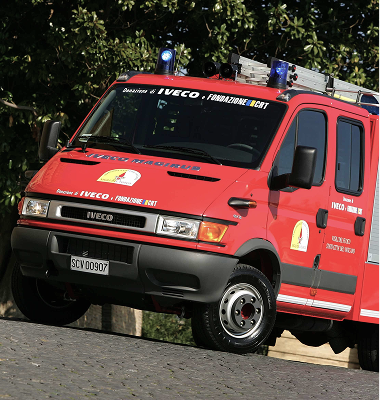 Red Iveco emergency vehicle with blue lights parked on a cobblestone surface under trees.
