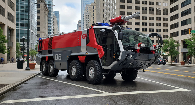 Large red and gray fire truck with multiple wheels parked on a city street amidst tall buildings.