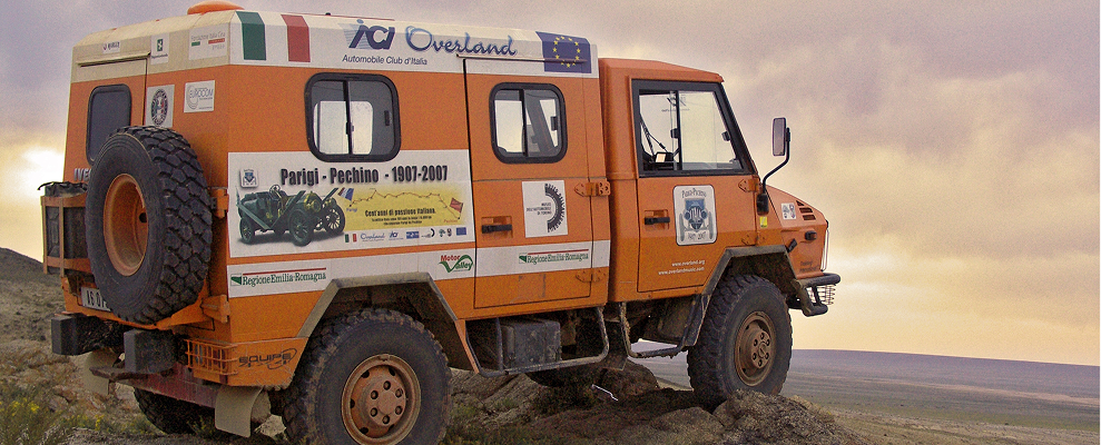 Orange expedition truck with Italian flags and logos parked on rocky terrain at sunset.