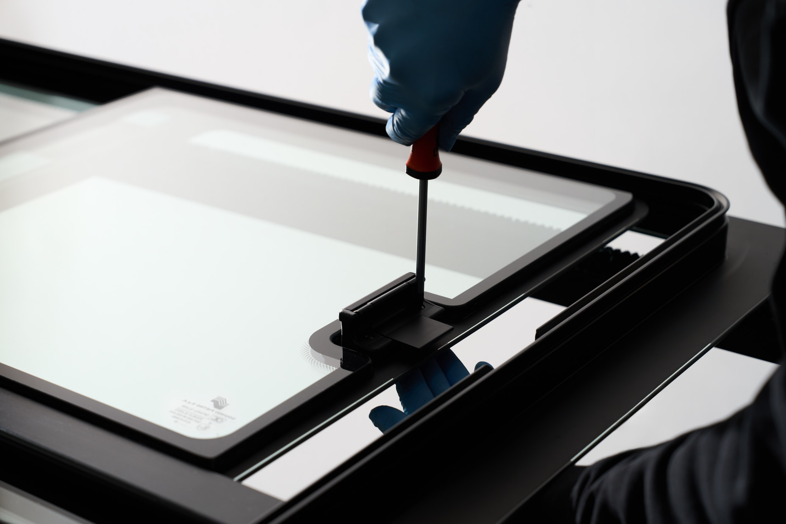 Person wearing blue gloves using screwdriver on black hinge attached to glass panel.