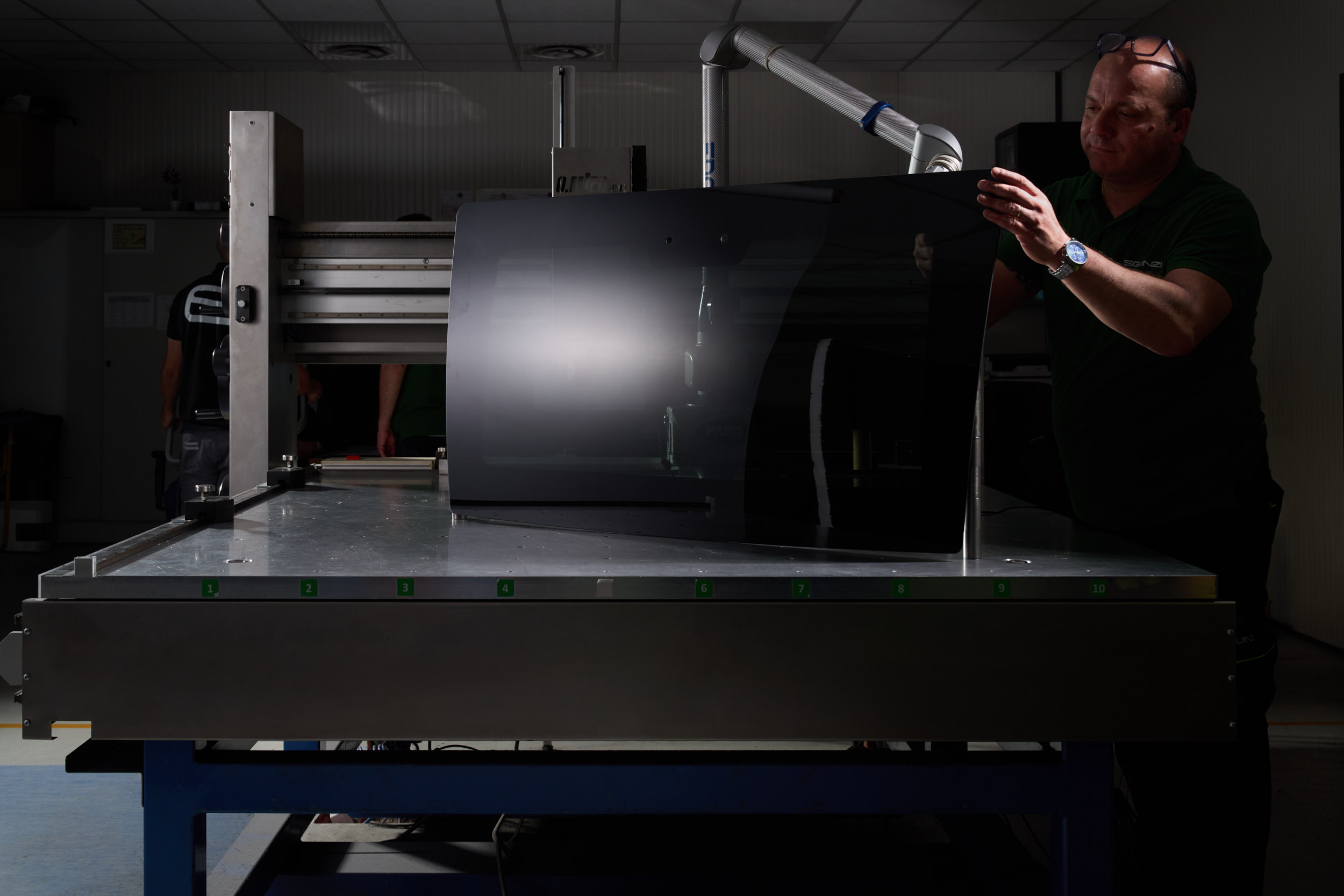 A man inspects a large glossy black screen panel on a metal industrial table in a factory setting.