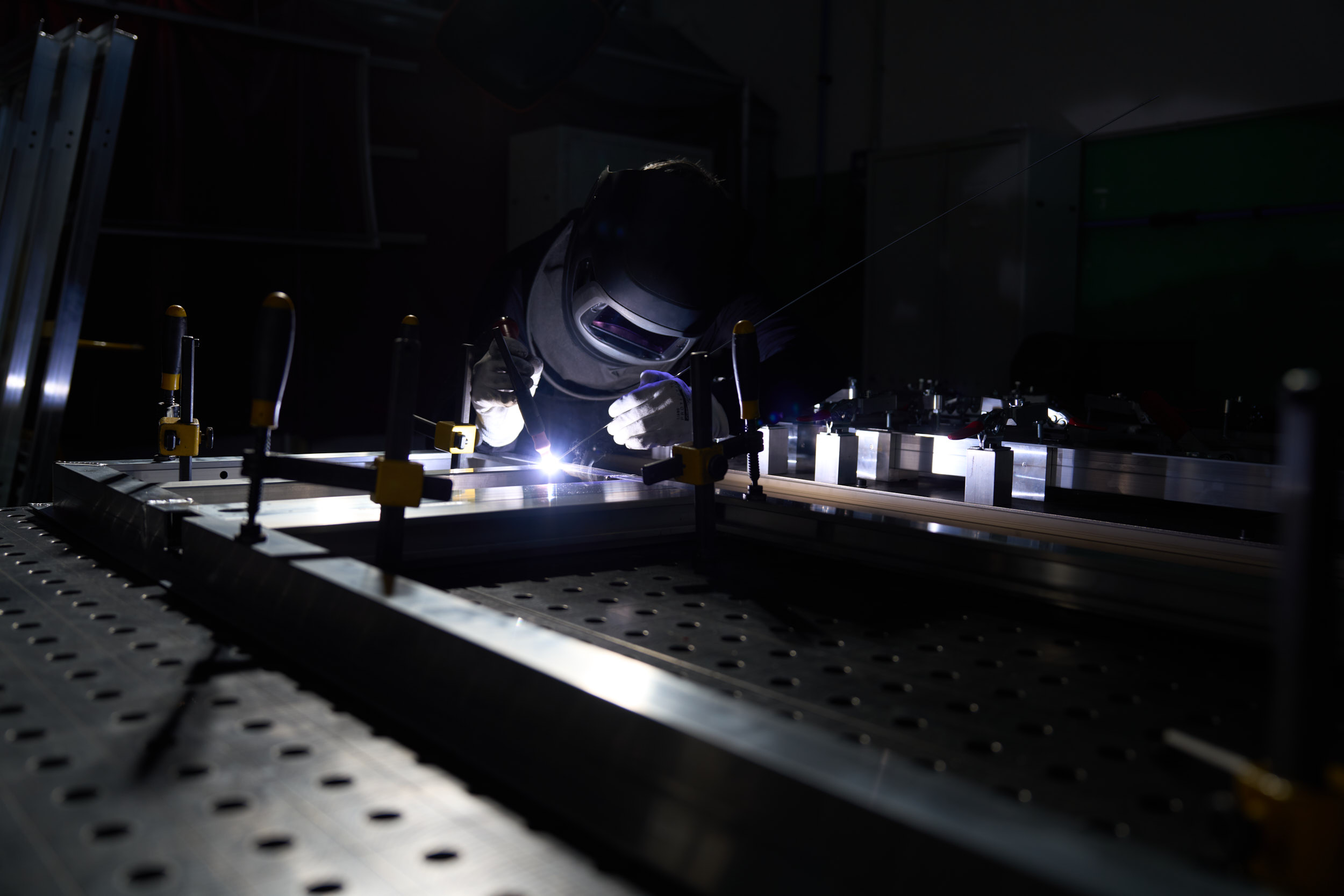 Welder wearing helmet and gloves working on metal frame in dimly lit workshop.