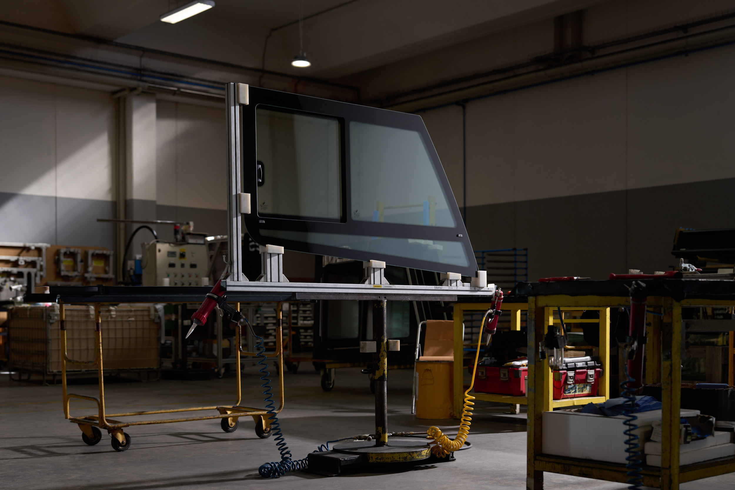 Car door frame held on a yellow metal stand inside a dimly lit industrial workshop.