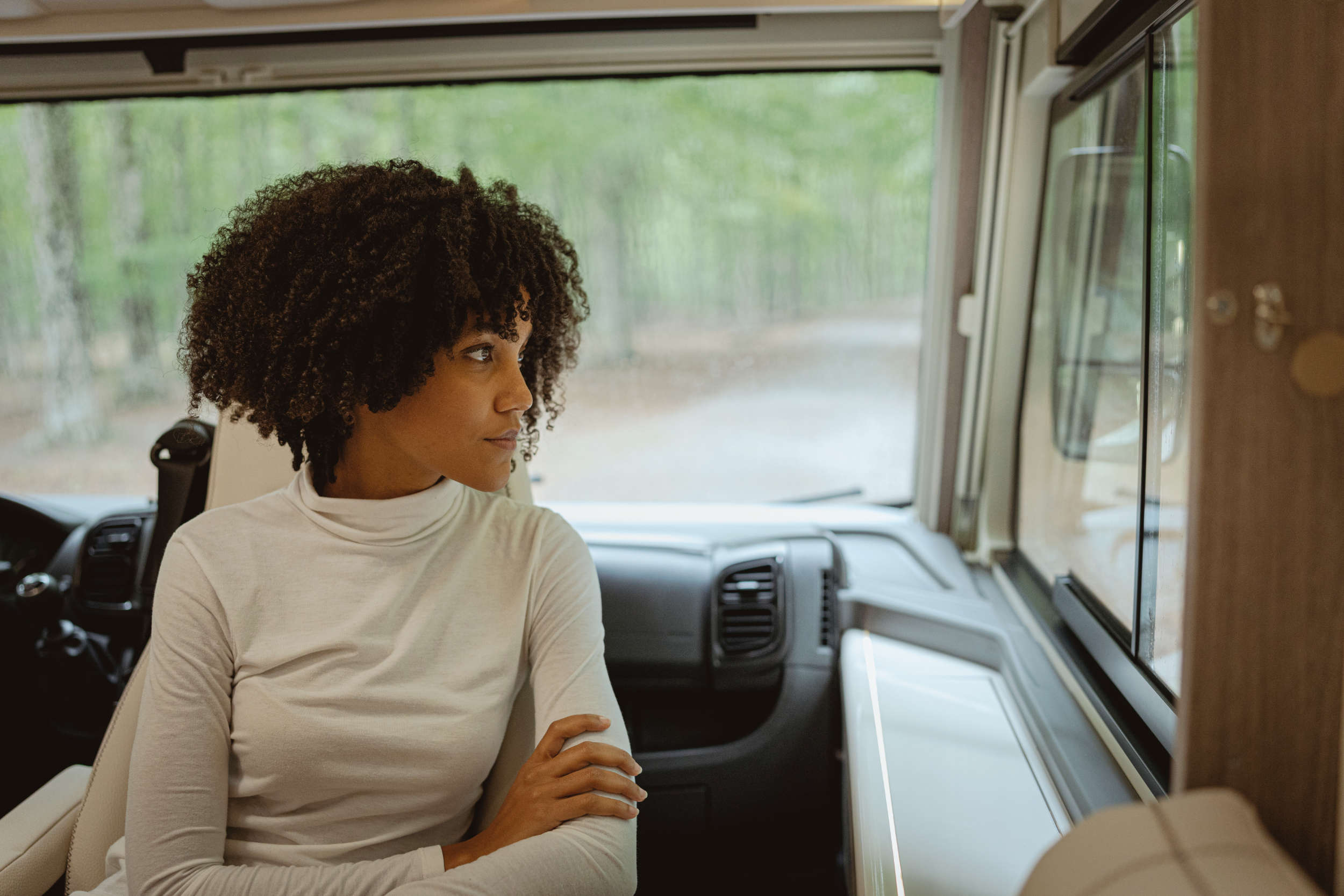 Woman with curly hair in white turtleneck looking out window inside a vehicle with forest outside.