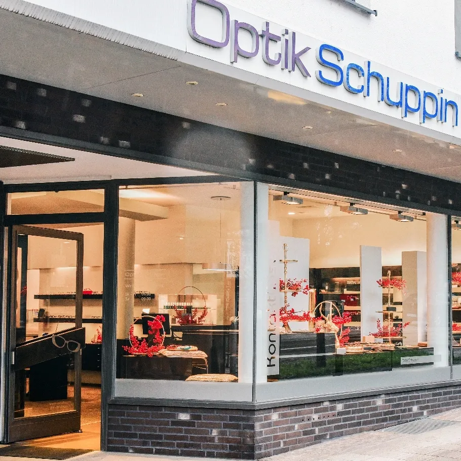 Außenansicht eines Optikergeschäfts mit großen Schaufenstern und roter Dekoration im Inneren.