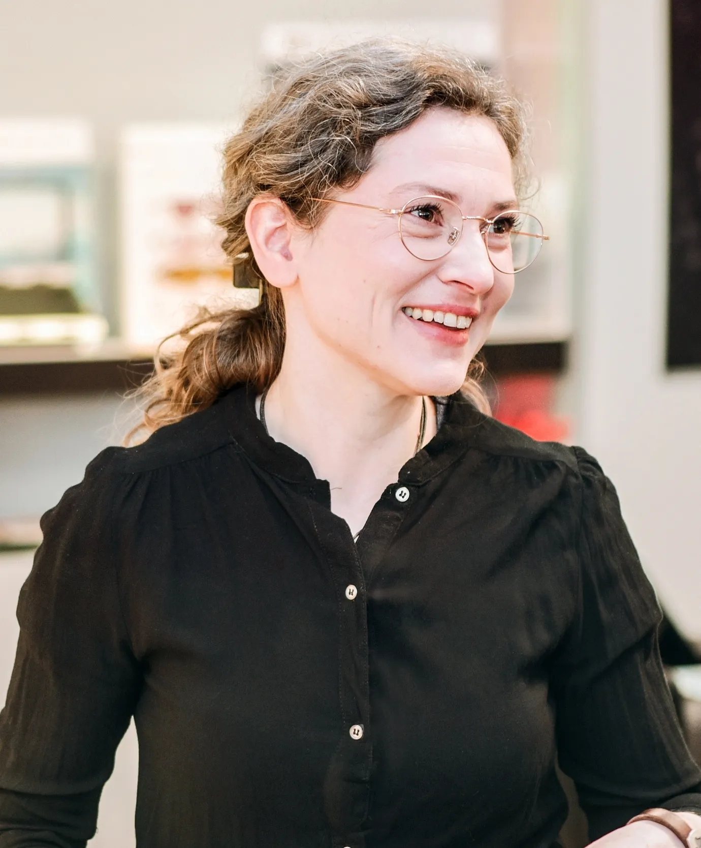 Frau mit Brille und braunem, lockigem Haar, in einem schwarzen Oberteil lächelt.