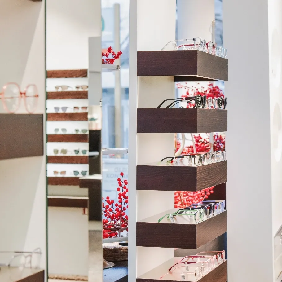 Mehrere Brillen auf dunklen Holzregalen in einem gut beleuchteten Geschäft mit Deko im Hintergrund.