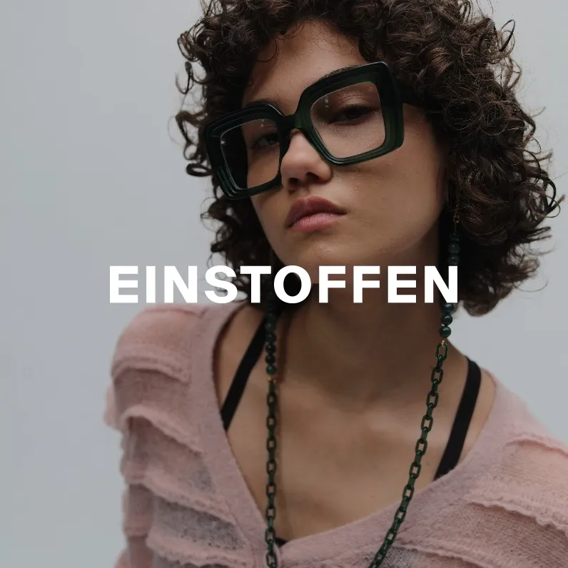 Markenbild Einstoffen. Porträt einer Frau mit lockigem Haar, die große, schwarze, quadratische Brille trägt und einen rosa Pullover anhat.