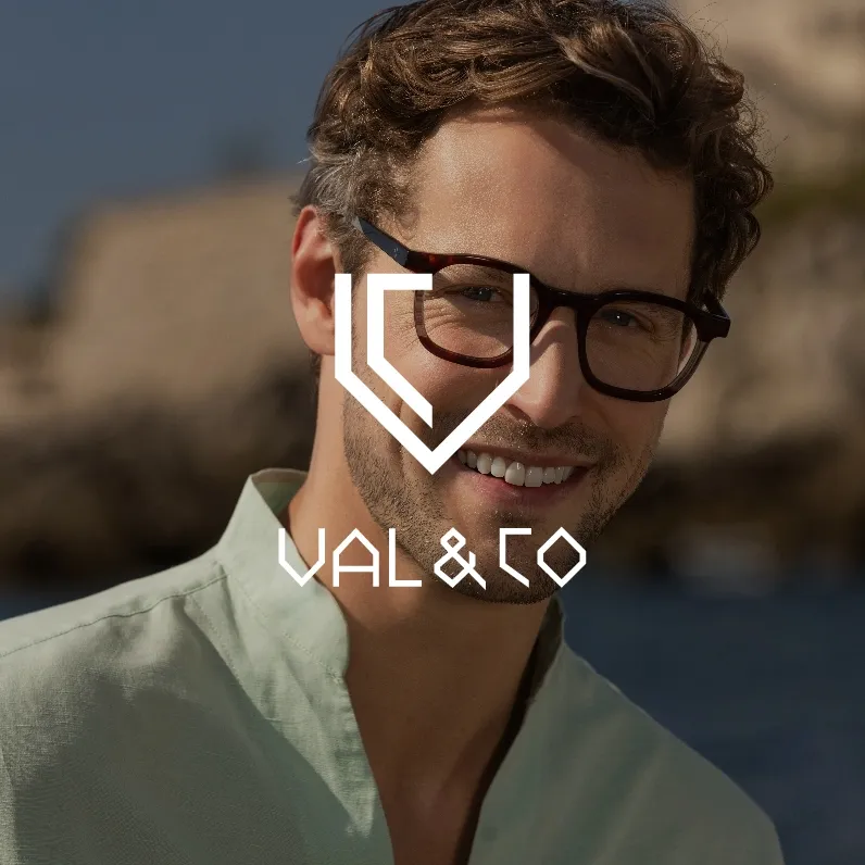 Markenbild Val & Co. Lächelnder Mann mit lockigem Haar und Brille vor verschwommenem Fels und Wasser im Hintergrund.