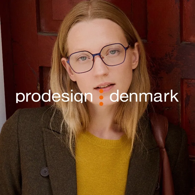Markenbild prodesign denmark. Porträt einer Frau mit blonden Haaren, die eine Brille trägt, in einem grünen Mantel und einem gelben Pullover vor einer roten Holztür.