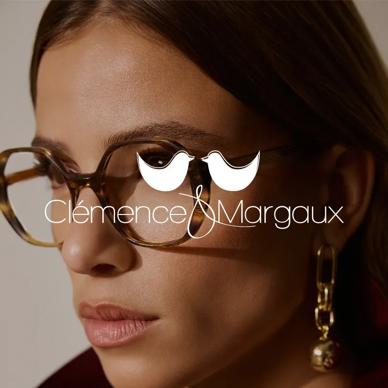 Clémence & Margaux. Nahaufnahme einer Frau mit braunen Haaren, die braune, rahmenlose Brille trägt und goldenen Ohrring mit Anhänger.