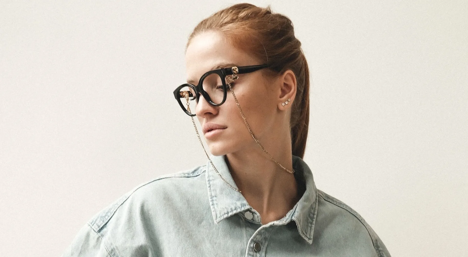 Frau mit roten Haaren, brauner Brille mit goldener Kette und grauem Jeanshemd vor hellem Hintergrund.