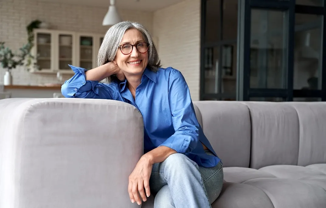 Fröhliche ältere Frau mit grauen Haaren und Brille sitzt entspannt auf einem grauen Sofa in einem hellen Wohnzimmer.